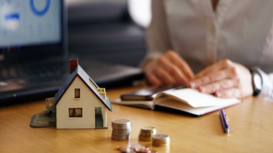 Imagen ilustrativa. Casa de juguete, monedas y persona haciendo cálculos